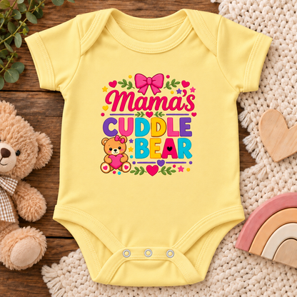 A yellow short-sleeve baby bodysuit laid flat on a wooden surface, surrounded by a plush teddy bear, a knit blanket, wooden hearts, and a pastel rainbow. The bodysuit features a colorful graphic that reads 'Mama's Cuddle Bear' with a pink bow, green leaves, and a small teddy bear illustration.