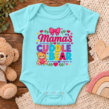 A light blue baby bodysuit with a colorful 'Mama's Cuddle Bear' graphic, styled in a flat lay on a wooden surface with a teddy bear, knit blanket, and wooden heart.