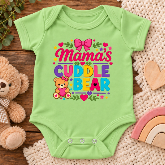 A light green baby bodysuit laid flat on a wooden surface, decorated with a colorful 'Mama's Cuddle Bear' graphic. The bodysuit is styled with a plush teddy bear, a wooden heart, a crochet blanket, and a small rainbow in the background.