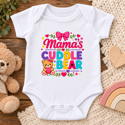 A white short-sleeve baby bodysuit laid flat on a wooden surface, surrounded by a plush teddy bear, a crochet blanket, and small wooden toys. The bodysuit has a colorful graphic that reads “Mama’s Cuddle Bear” with a pink bow, hearts, flowers, and a small teddy bear illustration.