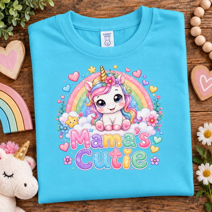 A folded aqua blue t-shirt with a colorful 'Mama's Cutie' unicorn graphic, displayed on a wooden surface with cute decorative props like a plush unicorn, wooden hearts, and flowers.