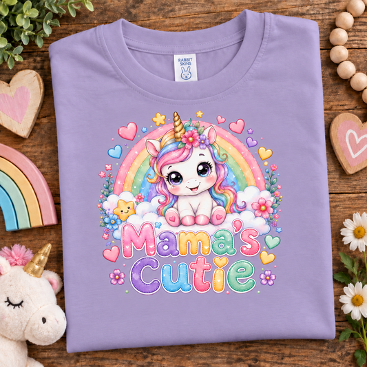A folded lavender crewneck t-shirt with a colorful cartoon unicorn graphic and the text 'Mama's Cutie' on the front, displayed on a wooden surface with cute decorative props.
