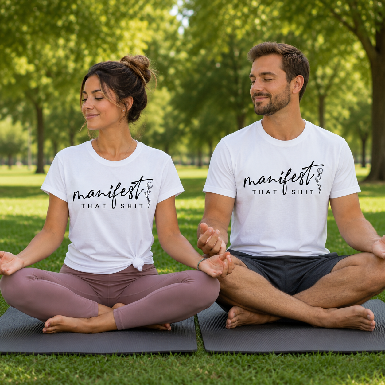 A man and a woman sitting cross-legged on yoga mats in a park, wearing matching white graphic t-shirts that read 'manifest THAT SHIT' in black script and block lettering.