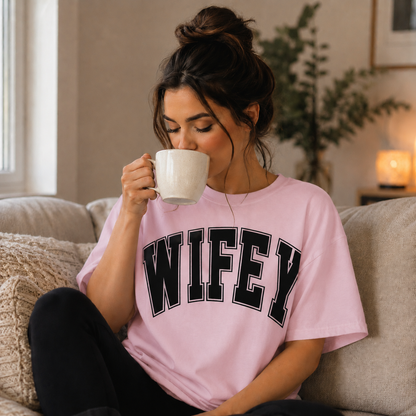 Woman wearing a 'WIFEY' light pink t-shirt, sitting on a couch and drinking from a mug.