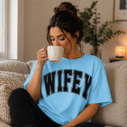 Woman wearing a 'WIFEY' sky blue colored t-shirt, sitting on a couch and drinking from a mug.