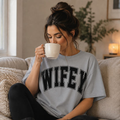 Woman wearing a 'WIFEY' sport gray colored t-shirt, sitting on a couch and drinking from a mug.