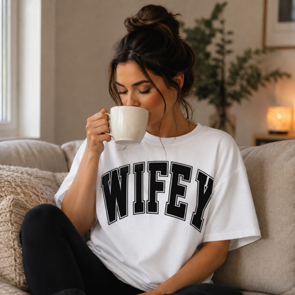 Woman wearing a 'WIFEY' white t-shirt, sitting on a couch and drinking from a mug.