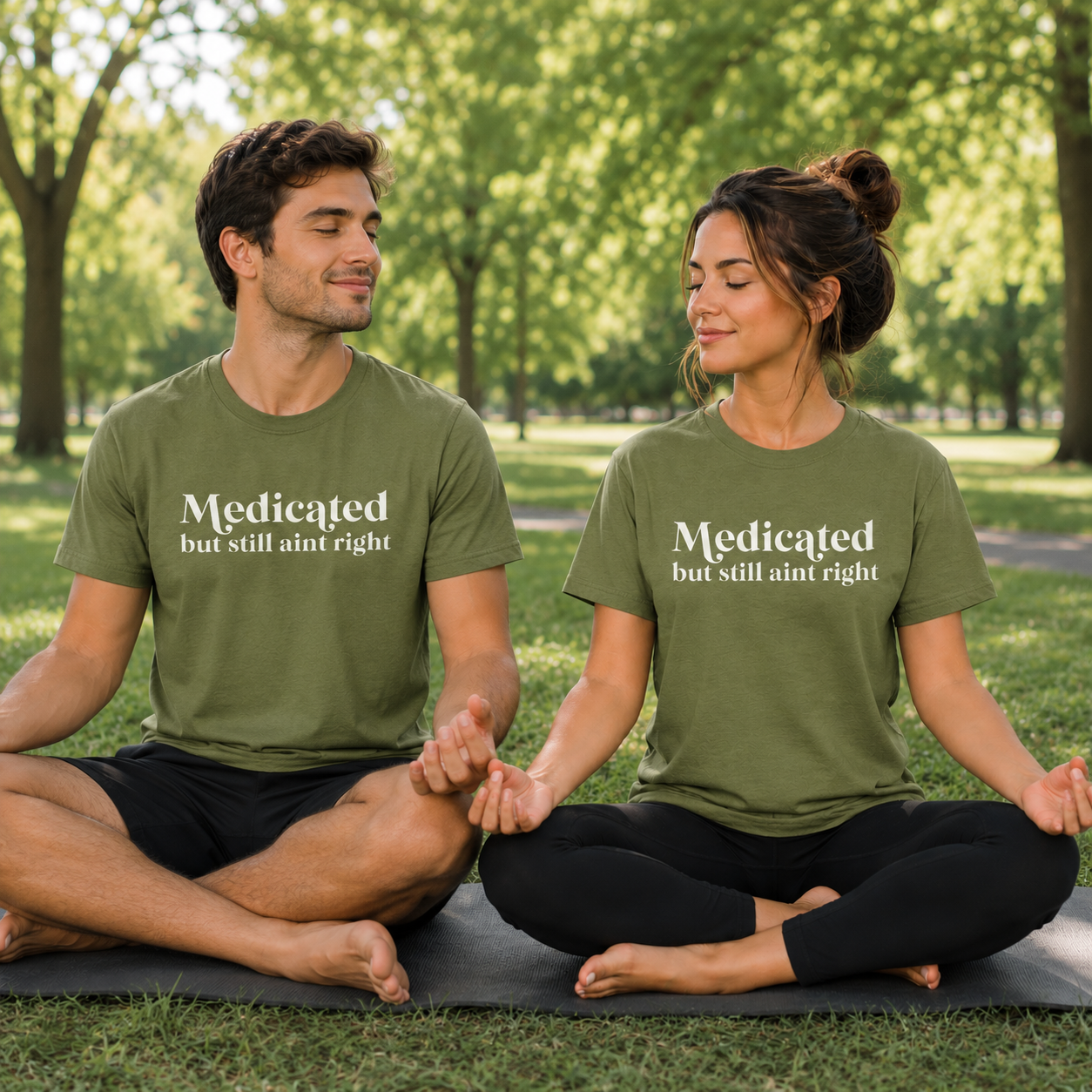 Two people sitting on yoga mats in a park wearing moss colored matching t-shirts with text Medicated but still ain't right.