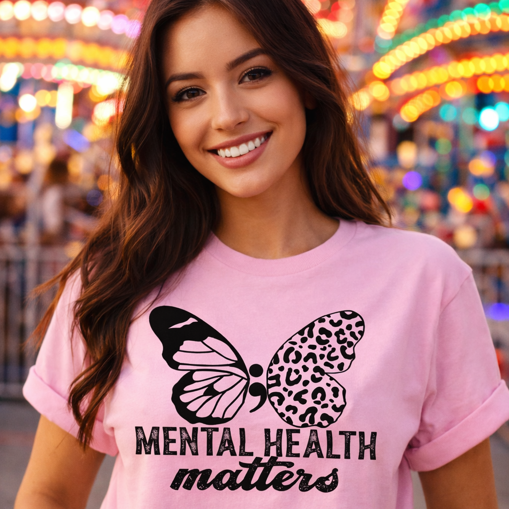 Woman wearing a pink t-shirt with a butterfly design and 'Mental Health Matters' text at a colorful fairground.