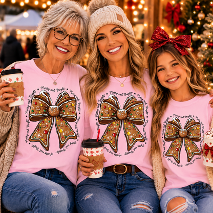 Three women posing together in matching light pink Christmas-themed t-shirts featuring a large glittery bow graphic with the text 'You are my happy holiday' in a circular design. The background shows a festive, warmly lit Christmas tree and holiday decorations.