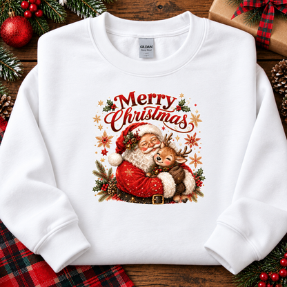 A white crewneck sweatshirt with a colorful Christmas graphic of Santa Claus and a reindeer under the text 'Merry Christmas'. The sweatshirt is displayed in a flat-lay setting on a rustic wooden background with holiday decorations like pine branches, ornaments, and a wrapped gift.