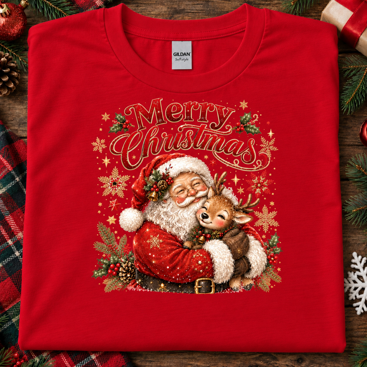 A folded red Gildan Softstyle Christmas t-shirt with a festive graphic of Santa Claus hugging a reindeer under the words 'Merry Christmas'. The shirt is styled on a rustic wooden background with holiday decorations like pine branches, a plaid blanket, and ornaments.