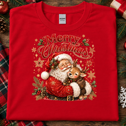 A folded red Gildan Softstyle Christmas t-shirt with a festive graphic of Santa Claus hugging a reindeer under the words 'Merry Christmas'. The shirt is styled on a rustic wooden background with holiday decorations like pine branches, a plaid blanket, and ornaments.