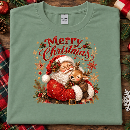 A folded sage green Gildan t-shirt with a festive Christmas graphic of Santa Claus hugging a reindeer under the text 'Merry Christmas'. The shirt is displayed on a rustic wooden background decorated with pine branches, a plaid blanket, and holiday ornaments.