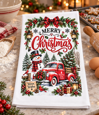 A white kitchen tea towel with a festive Christmas print, featuring a red vintage truck, snowman, pine trees, and the words 'Merry Christmas' in red script, styled on a marble countertop with baking props.