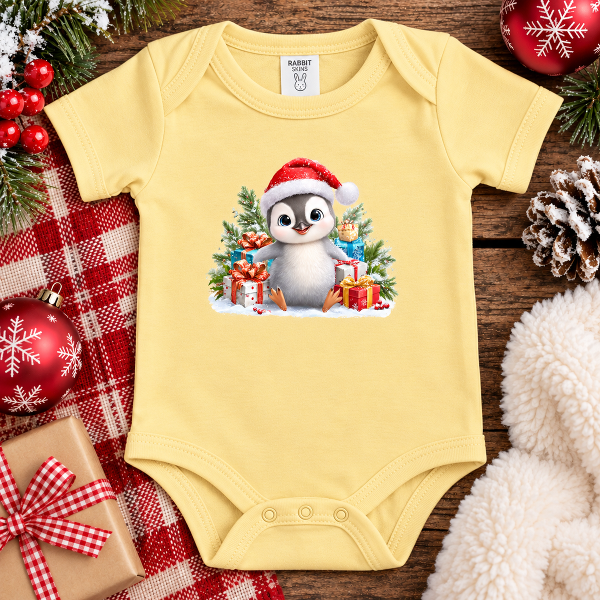 A butter yellow baby bodysuit with a Christmas penguin graphic, styled in a flat lay on a wooden surface with holiday decorations including a red plaid blanket, ornaments, pinecones, and a white faux fur blanket.