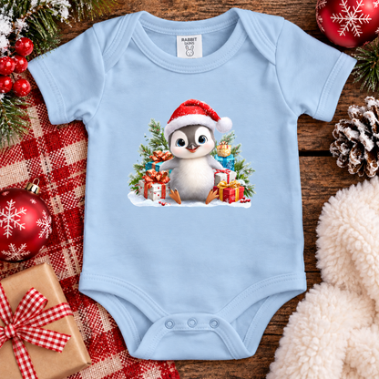 A light blue baby bodysuit with a festive Christmas penguin graphic, styled in a holiday flat-lay with red plaid fabric, pine branches, red ornaments, and a soft cream faux-fur texture.