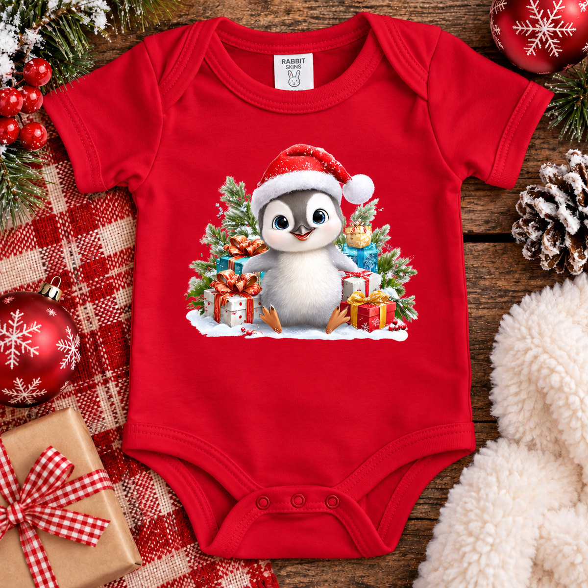 A red short-sleeve baby bodysuit laid flat on a rustic wooden surface, decorated with a large Christmas penguin graphic wearing a Santa hat and surrounded by gifts and evergreen trees. The bodysuit is styled with holiday ornaments, a red-and-white plaid blanket, pinecones, and a cream faux-fur textile.
