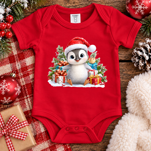 A red short-sleeve baby bodysuit laid flat on a rustic wooden surface, decorated with a large Christmas penguin graphic wearing a Santa hat and surrounded by gifts and evergreen trees. The bodysuit is styled with holiday ornaments, a red-and-white plaid blanket, pinecones, and a cream faux-fur textile.