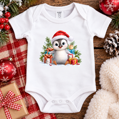 A white baby bodysuit laid flat on a rustic wooden surface, decorated with a festive Christmas graphic of a penguin in a Santa hat surrounded by gifts and evergreen trees. The bodysuit is styled with holiday props including red ornaments, a plaid blanket, pinecones, and a fluffy cream blanket.