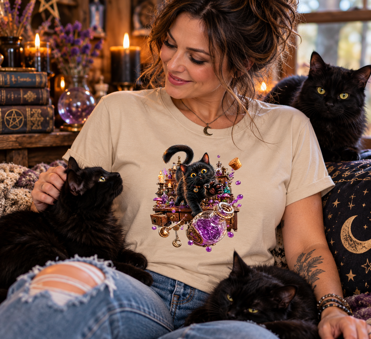 A woman wearing a sand graphic t-shirt with a black cat and purple crystals design, sitting with three black cats in a cozy, candle-lit room.