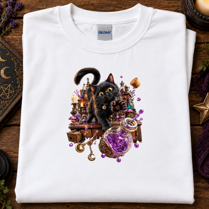 A folded white Gildan t-shirt displayed flat on a rustic wooden surface, featuring a detailed graphic print of a black cat surrounded by purple crystals, a cauldron, and magical props.