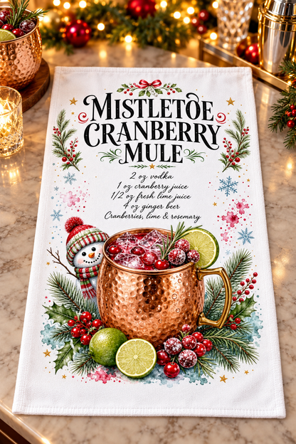 A white kitchen towel featuring a festive 'Mistletoe Cranberry Mule' recipe and graphic design laid flat on a marble countertop with holiday lights and copper mugs in the background.
