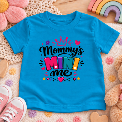 A turquoise blue short-sleeve kids' T-shirt laid flat on a soft cream rug with pastel toys around it. The shirt has a colorful graphic reading “Mommy’s Mini Me” with a crown, hearts, stars, and multicolored lettering.