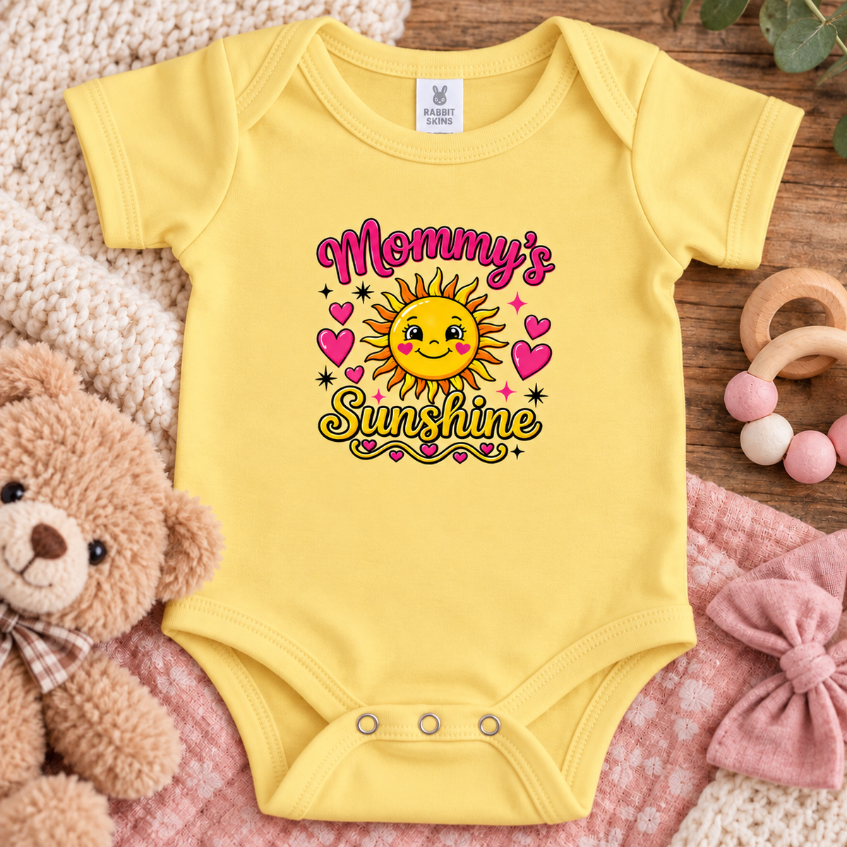 A butter short-sleeve baby bodysuit laid flat on a pink blanket, surrounded by a teddy bear, knit blanket, wooden toy, and pink bow. The bodysuit has a colorful graphic that reads 'Mommy's Sunshine' with a smiling sun and pink hearts.