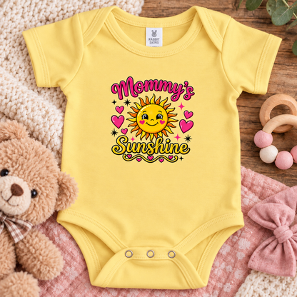 A butter short-sleeve baby bodysuit laid flat on a pink blanket, surrounded by a teddy bear, knit blanket, wooden toy, and pink bow. The bodysuit has a colorful graphic that reads 'Mommy's Sunshine' with a smiling sun and pink hearts.