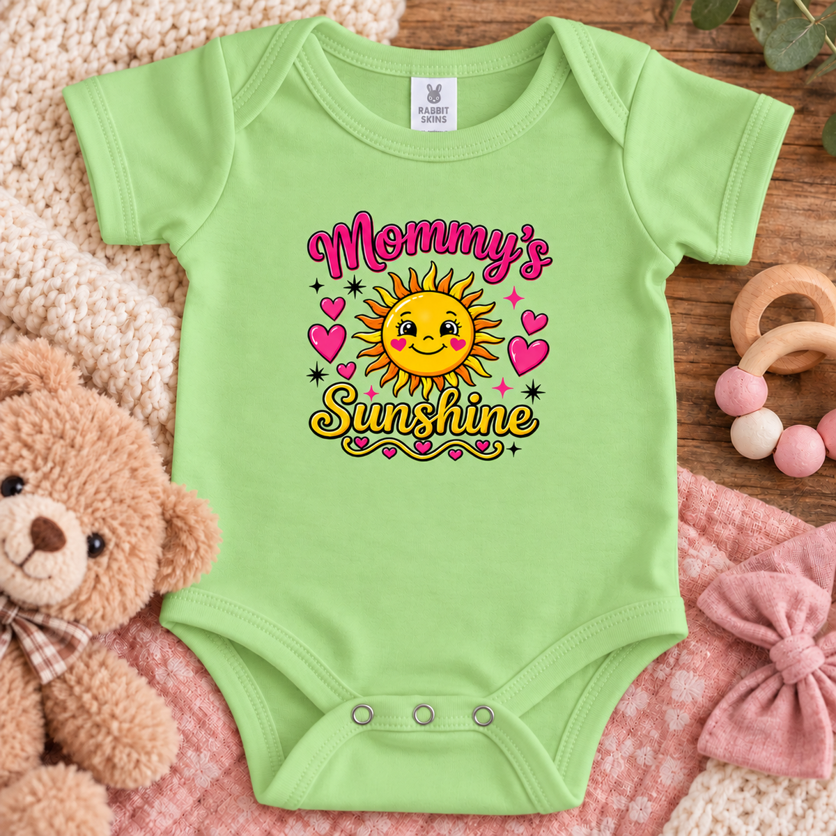 A keylime green baby bodysuit laid flat on a pink blanket with a teddy bear, wooden beads, and a knitted textile around it. The bodysuit has a colorful graphic that reads 'Mommy's Sunshine' with a smiling sun, hearts, and stars.