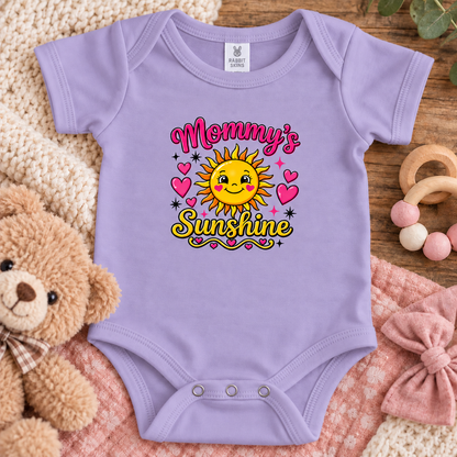 A lavender baby bodysuit with a colorful 'Mommy's Sunshine' graphic, styled in a cozy flat-lay arrangement with a teddy bear, knit blankets, and wooden toys.