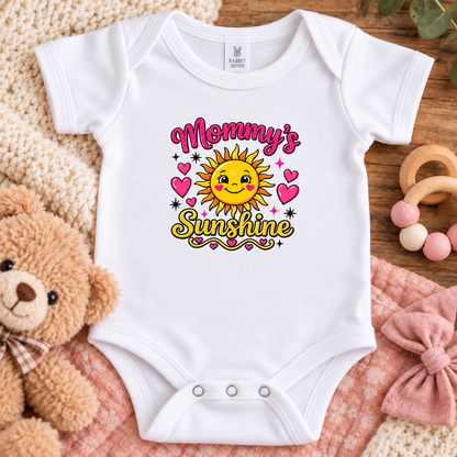 A white short-sleeve baby bodysuit laid flat on a pink blanket over a wooden surface, surrounded by a teddy bear, knit blanket, wooden toy, and pink bow. The bodysuit has a colorful graphic reading 'Mommy's Sunshine' with a smiling sun and pink hearts.
