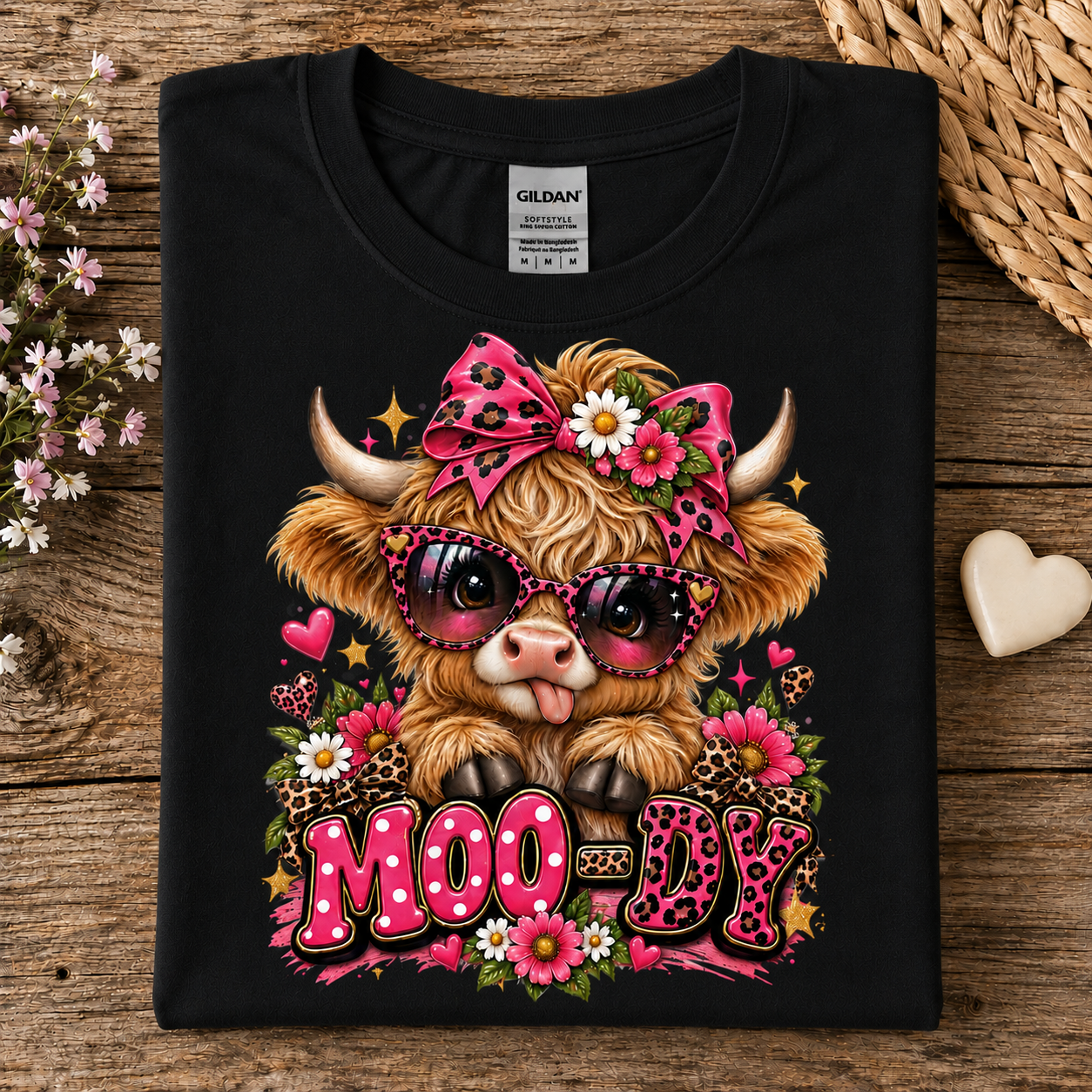 A black Gildan t-shirt laid flat on a rustic wooden surface, featuring a colorful graphic of a fluffy Highland cow wearing pink leopard-print sunglasses and a matching bow, surrounded by flowers and the word 'MOODY' in pink leopard-print letters.