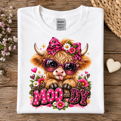 A folded white Gildan t-shirt displayed flat on a rustic wooden surface, featuring a colorful graphic of a fluffy Highland cow wearing pink leopard-print sunglasses and a matching bow, surrounded by flowers and the text 'MOODY'.