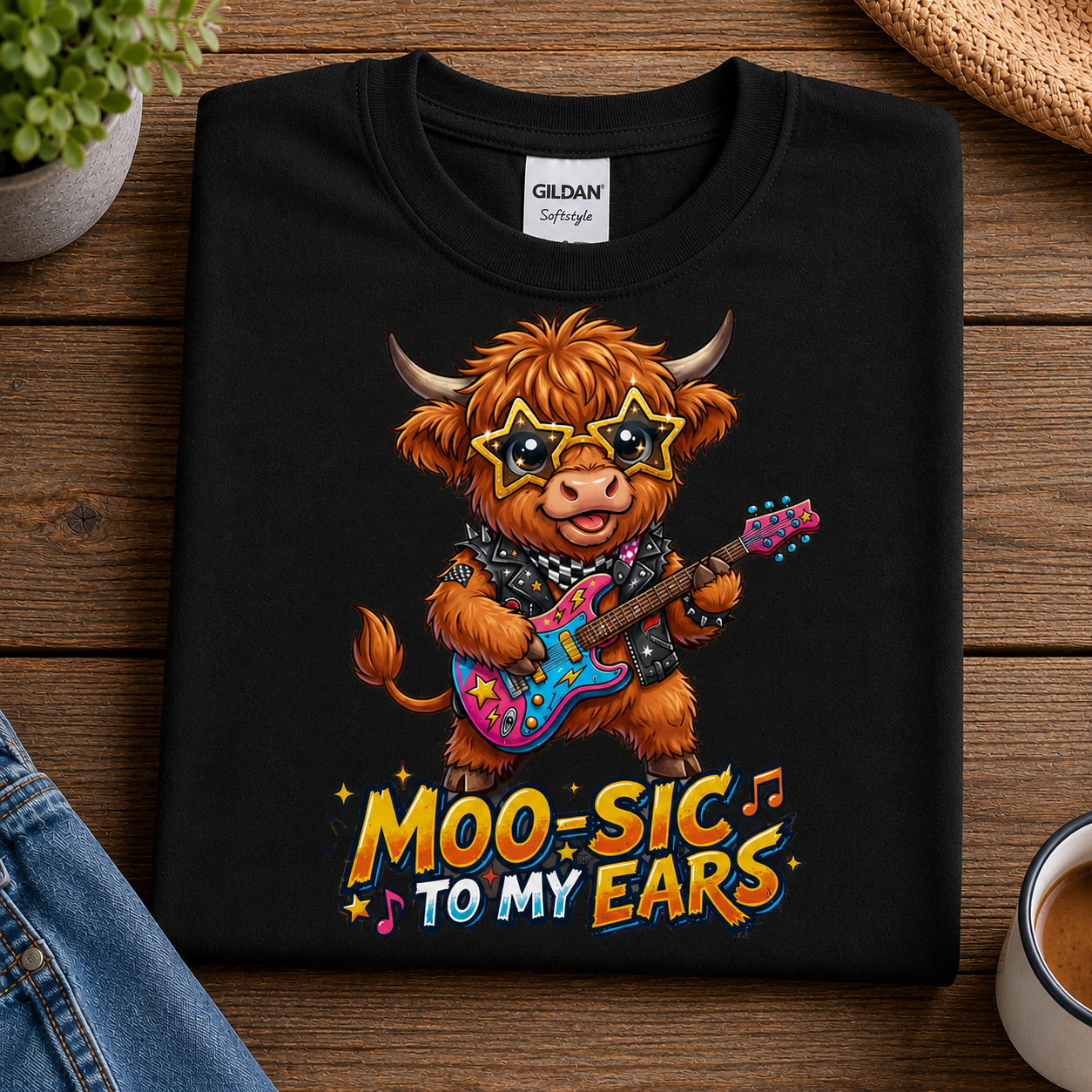 A black Gildan Softstyle t-shirt laid flat on a wooden surface, featuring a cartoon Highland cow playing a colorful guitar with the text 'MOO-SIC TO MY EARS'.
