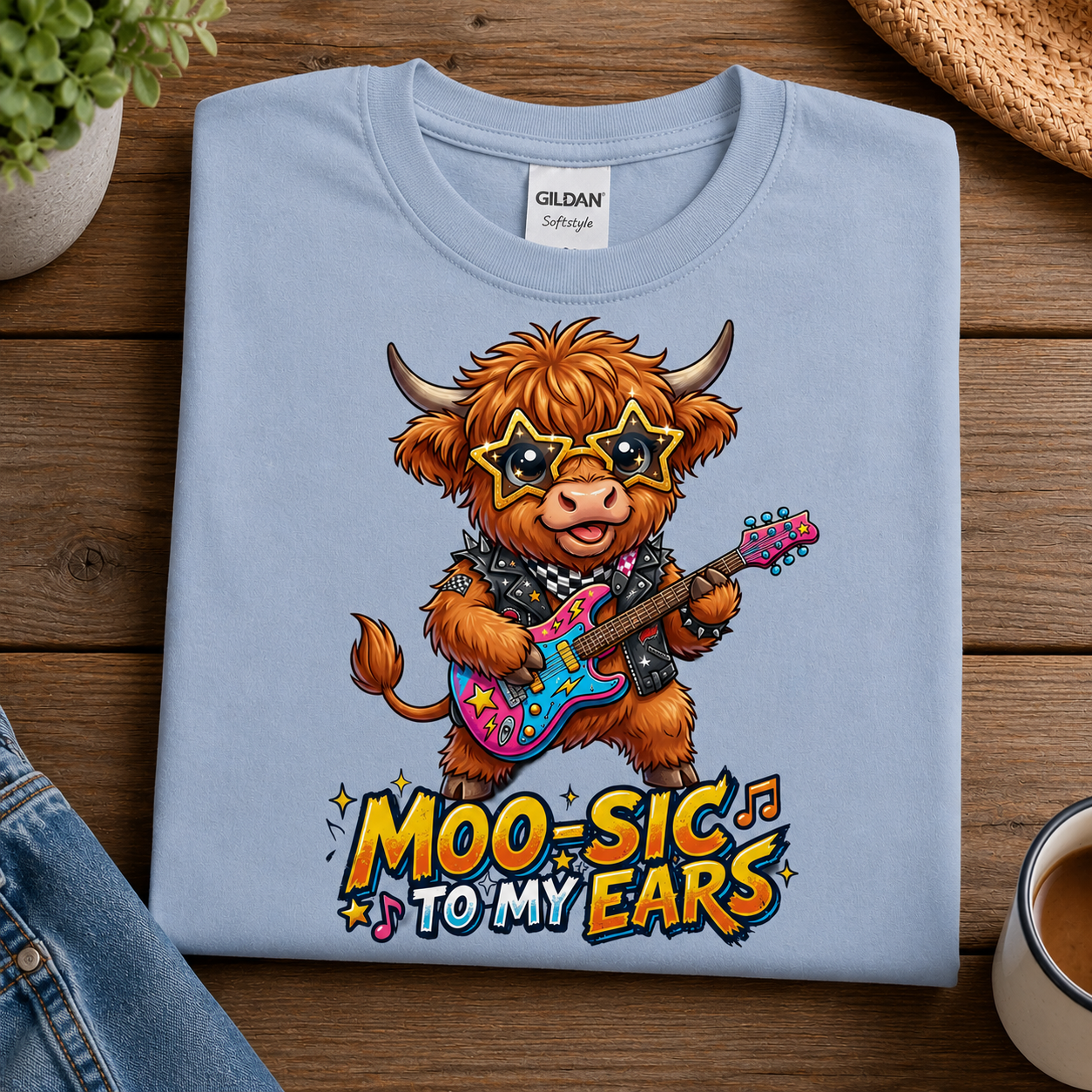 A light blue Gildan Softray tees folded t-shirt with a colorful graphic of a rock-crazy Highland cow playing an electric guitar, with the text 'MOO-SIC TO MY EARS' below it. The t-shirt is displayed on a wooden table with lifestyle props including a plant, denim, and a coffee cup.