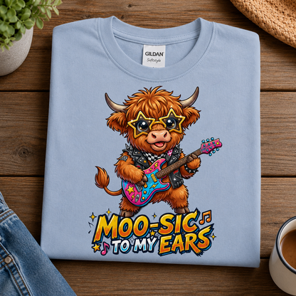 A light blue Gildan Softray tees folded t-shirt with a colorful graphic of a rock-crazy Highland cow playing an electric guitar, with the text 'MOO-SIC TO MY EARS' below it. The t-shirt is displayed on a wooden table with lifestyle props including a plant, denim, and a coffee cup.