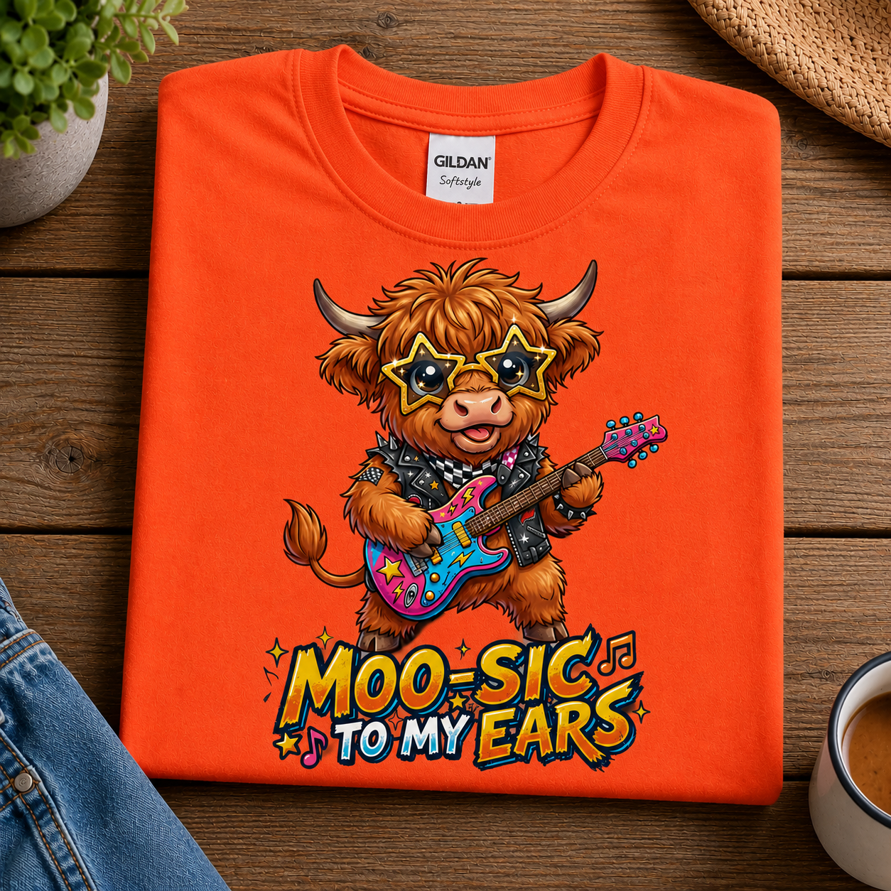 An orange Gildan t-shirt is neatly folded on a rustic wooden surface, featuring a cartoon Highland cow playing a colorful guitar with the text 'MOO-SIC TO MY EARS'.