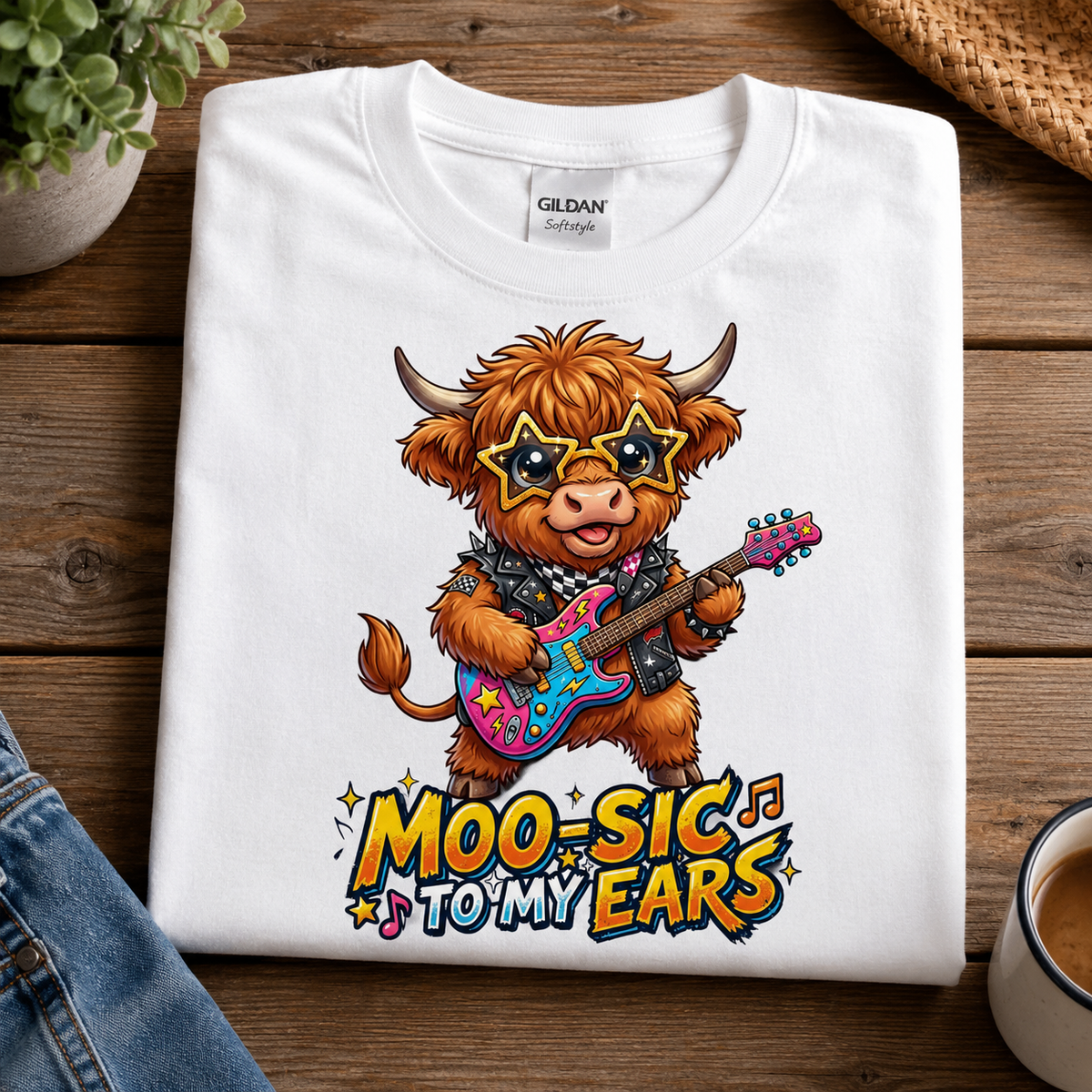 A folded white Gildan t-shirt with a colorful cartoon graphic of a fluffy brown cow playing a pink and blue electric guitar, with the text 'MOO-SIC TO MY EARS' below. The shirt is displayed on a wooden table flat-lay with a plant, denim, and a coffee cup nearby.