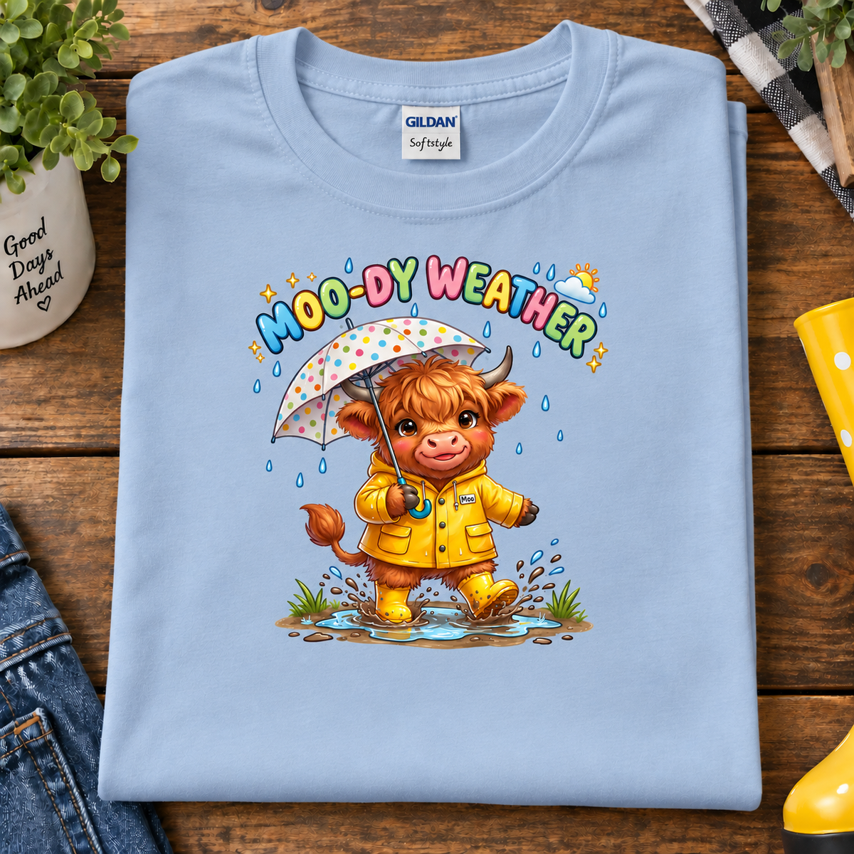 A folded light blue Gildan Sofstyle t-shirt with a colorful cartoon graphic of a cow in a yellow raincoat holding a polka-dot umbrella under the text 'MOO-DY WEATHER'. The shirt is styled in a flat-lay on a wooden table with jeans, a plant, and a yellow rain boot.