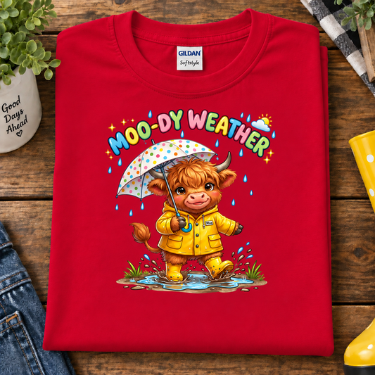 A folded red Gildan Softstyle T-shirt with a colorful cartoon graphic of a cow in a yellow raincoat holding a polka-dot umbrella under the text 'MOO-DY WEATHER,' styled on a wooden surface with jeans, a plant, and rain boots nearby.