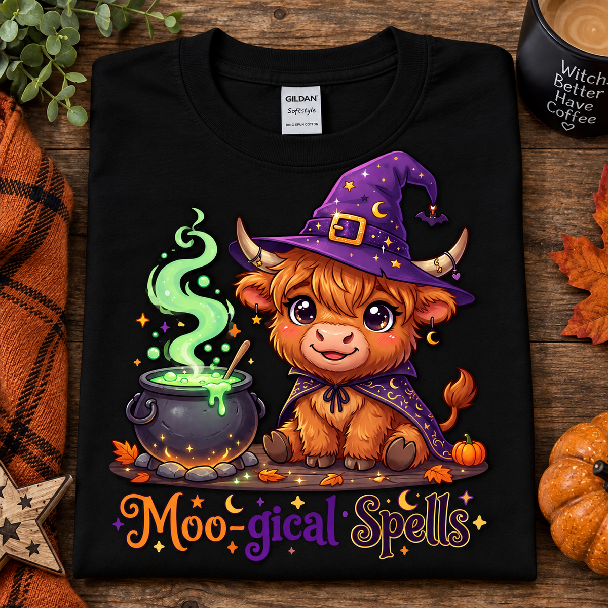 A folded black Gildan Comfort T-shirt with a colorful cartoon graphic of a fluffy brown creature wearing a purple witch hat and cape beside a cauldron, with the text 'Moo-gical Spells' below. The shirt is styled on a rustic wood surface with autumn leaves, a candle, and a small pumpkin.