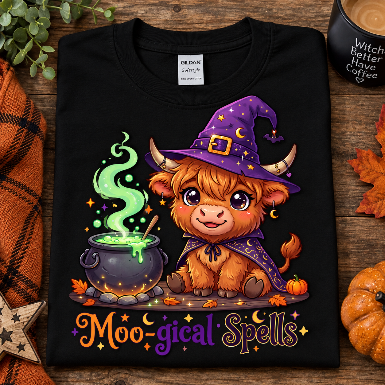 A folded black Gildan Comfort T-shirt with a colorful cartoon graphic of a fluffy brown creature wearing a purple witch hat and cape beside a cauldron, with the text 'Moo-gical Spells' below. The shirt is styled on a rustic wood surface with autumn leaves, a candle, and a small pumpkin.