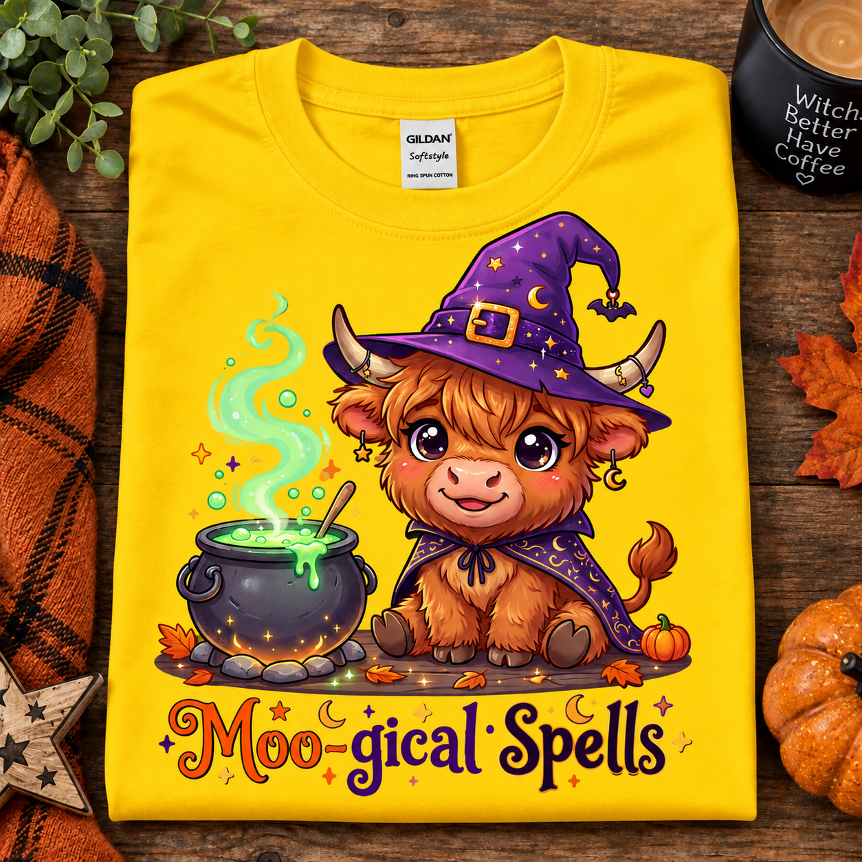 A folded daisy yellow Gildan Comfort T-shirt with a colorful cartoon graphic of a fluffy brown creature wearing a purple witch hat and cape beside a cauldron, with the text 'Moo-gical Spells' below. The shirt is styled on a rustic wood surface with autumn leaves, a candle, and a small pumpkin.