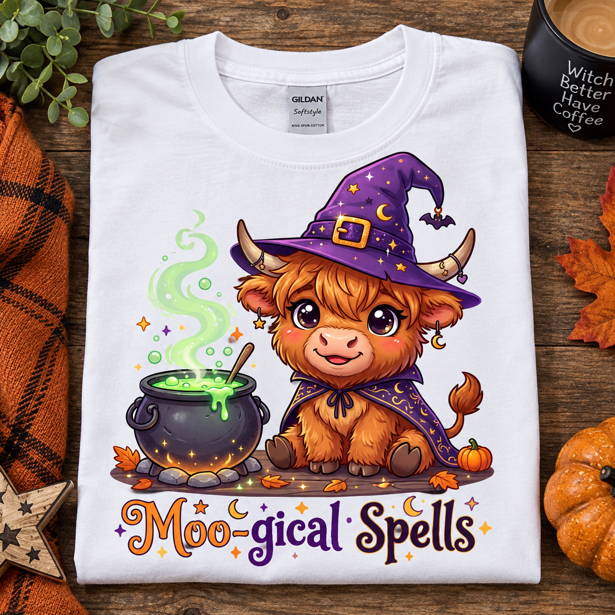 A white Gildan Softstyle T-shirt laid flat on a rustic wooden surface, featuring a cute cartoon witch goat character in a purple hat and cape beside a cauldron with green potion and the text 'Moo-gical Spells.' Decorative autumn props, a plaid blanket, leaves, and a mug are arranged around the shirt.