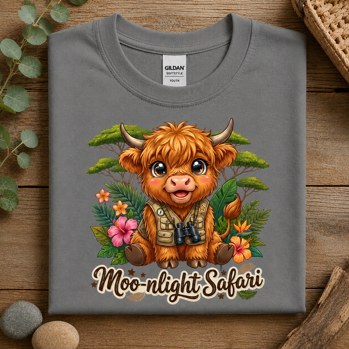 A folded heather graphite Gildan Comfort Colors T-shirt with a colorful cartoon Highland cow graphic and the text 'Moonlight Safari' on the front, displayed on a rustic wooden surface with decorative leaves and stones.