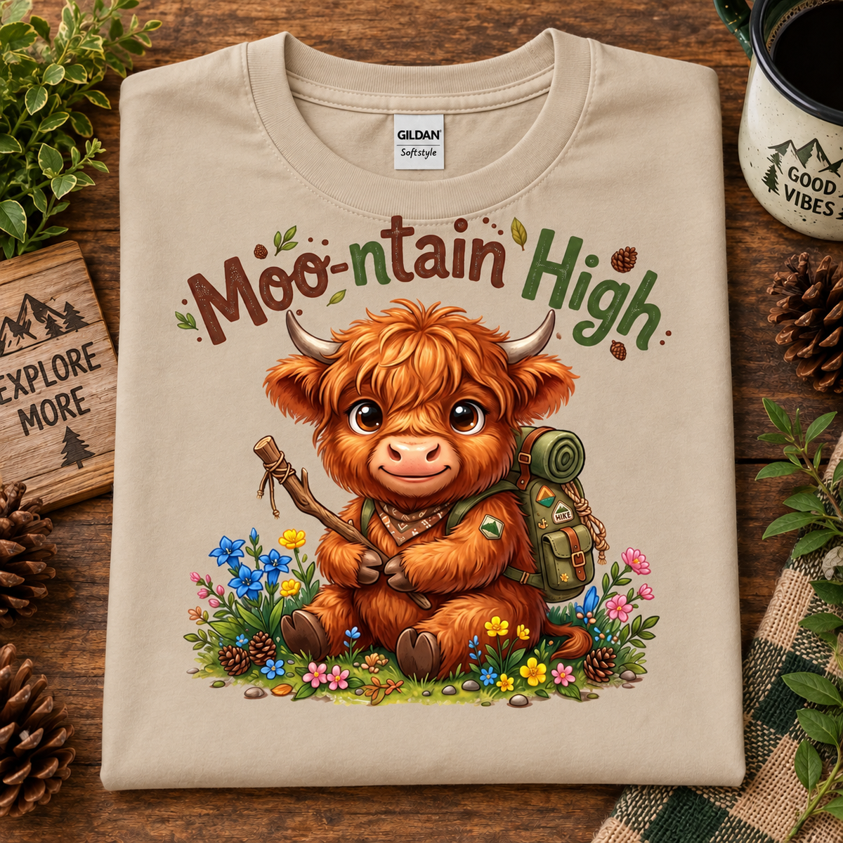 A sand colored folded t-shirt with a colorful graphic of a cute Highland cow holding a stick in a field of flowers, with the text 'Moo-ntain High' above it. The shirt is displayed on a rustic wooden background with pinecones, a mug, and greenery.