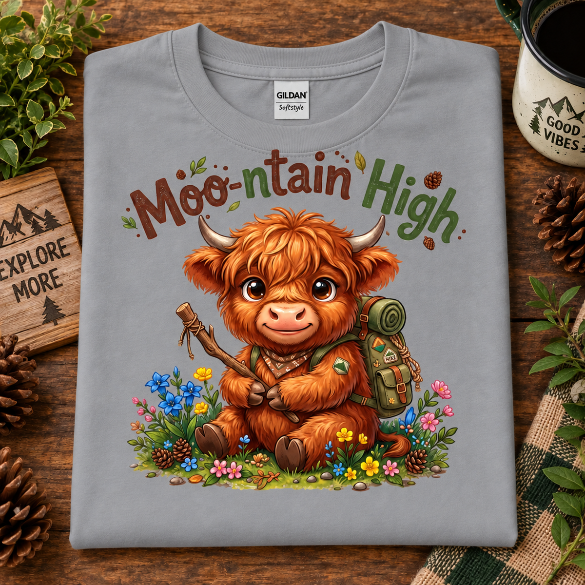 A folded sport gray Gildan Softstyle t-shirt with a colorful graphic of a cute Highland cow holding a stick in a flower field, with the text 'Moo-nountain High' above it. The shirt is displayed on a rustic wooden background with pinecones, a mug, and greenery.