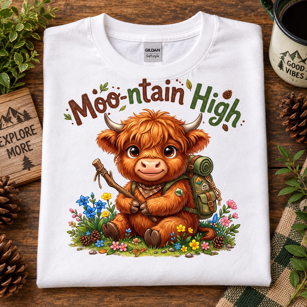 A folded white Gildan Softstyle t-shirt is displayed on a rustic wooden surface. The shirt features a colorful graphic of a cute, fluffy brown Highland cow wearing a green hiking backpack and holding a stick, surrounded by wildflowers, with the text 'Moo-ntain High' above it.